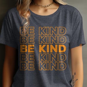 May include: Dark gray t-shirt with the words "BE KIND" repeated vertically in orange block letters. The shirt has a crew neck and short sleeves. The text is outlined in a darker orange.