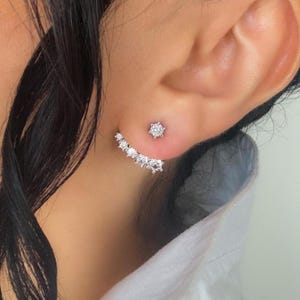 May include: A silver-toned ear jacket earring featuring a single round clear stone stud at the front and a curved, multi-stone ear crawler piece that sits below the earlobe, accented with five smaller round clear stones in a row.