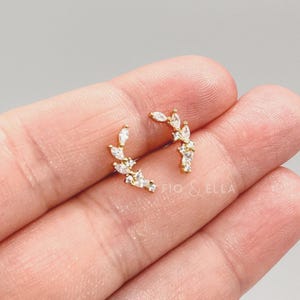 May include: A pair of gold-toned ear climbers featuring a curved, branch-like design adorned with multiple marquise and round-cut clear white crystals. The jewelry pieces are arranged in a delicate, ascending formation, showcasing a polished metallic finish.