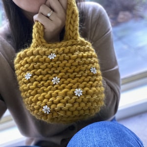 K&ouml;nnte beinhalten: Eine kleine, senfgelbe Stricktasche mit einem dicken Strickgriff und wei&szlig;en Blumenverzierungen.