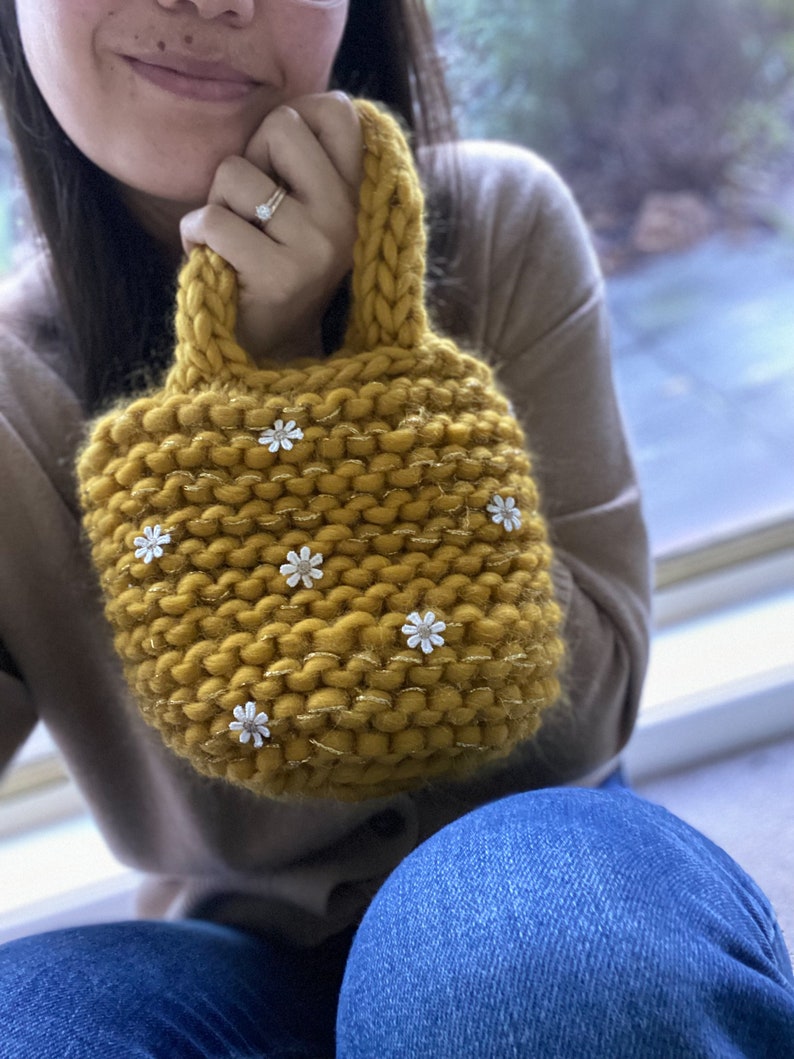 K&ouml;nnte beinhalten: Eine kleine, senfgelbe Stricktasche mit einem dicken Strickgriff und wei&szlig;en Blumenverzierungen.