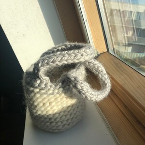 K&ouml;nnte beinhalten: Eine kleine, wei&szlig;e und graue Stricktasche mit zwei Henkeln. Die Tasche ist aus weichem, flauschigem Garn.