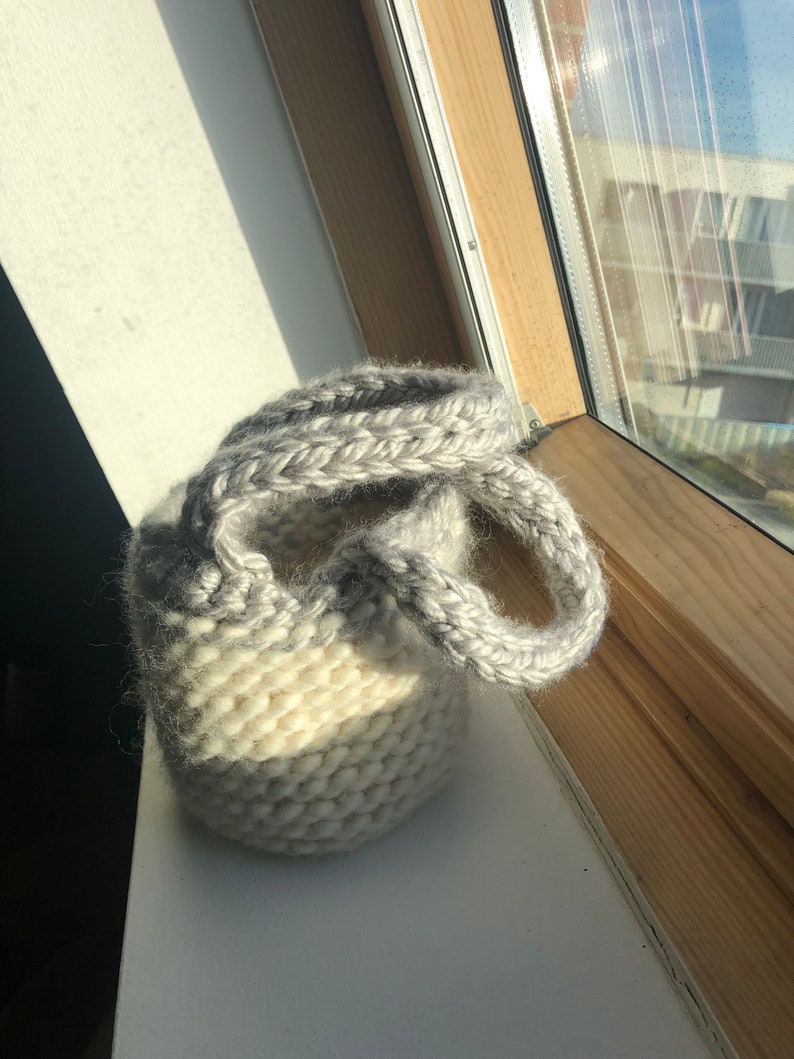 K&ouml;nnte beinhalten: Eine kleine, wei&szlig;e und graue Stricktasche mit zwei Henkeln. Die Tasche ist aus weichem, flauschigem Garn.