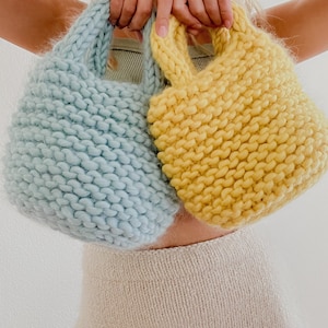K&ouml;nnte beinhalten: Zwei gestrickte Einkaufstaschen, eine hellblau und eine gelb, die von den H&auml;nden einer Person gehalten werden.