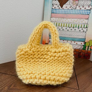 K&ouml;nnte beinhalten: Eine kleine, gelbe Stricktasche mit einem einzigen Henkel. Die Tasche ist aus dickem, grobem Garn gefertigt und hat eine leicht strukturierte Oberfl&auml;che.