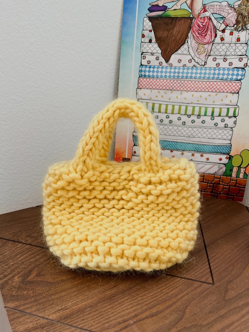 K&ouml;nnte beinhalten: Eine kleine, gelbe Stricktasche mit einem einzigen Henkel. Die Tasche ist aus dickem, grobem Garn gefertigt und hat eine leicht strukturierte Oberfl&auml;che.