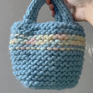 K&ouml;nnte beinhalten: Eine hellblaue gestrickte Tragetasche mit einem bunten Streifenband um den oberen Rand.