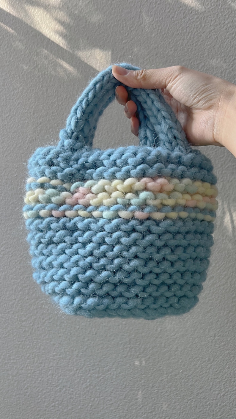 K&ouml;nnte beinhalten: Eine hellblaue gestrickte Tragetasche mit einem bunten Streifenband um den oberen Rand.