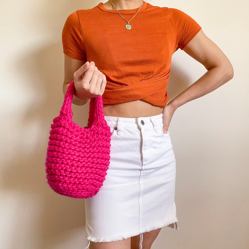 K&ouml;nnte beinhalten: Eine leuchtend pinkfarbene gestrickte Tragetasche mit einem kurzen wei&szlig;en Jeansrock und einem rostfarbenen T-Shirt.
