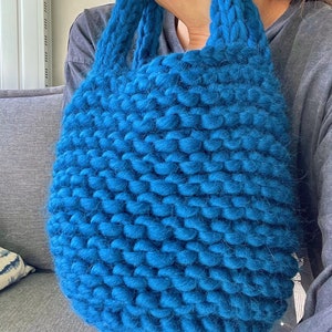 K&ouml;nnte beinhalten: Eine gro&szlig;e, tealblaue Stricktasche aus dickem, grobem Garn. Die Tasche hat eine breite, offene Oberseite und zwei dicke, gekn&uuml;pfte Henkel.