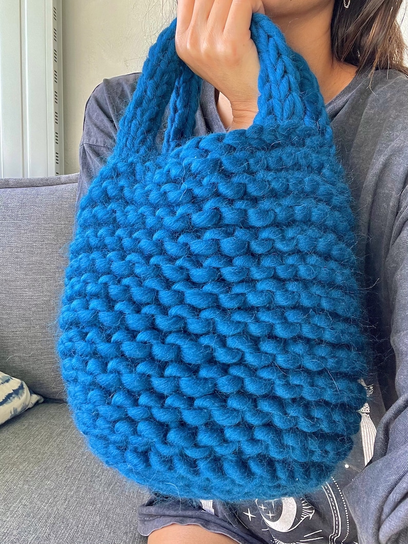 K&ouml;nnte beinhalten: Eine gro&szlig;e, tealblaue Stricktasche aus dickem, grobem Garn. Die Tasche hat eine breite, offene Oberseite und zwei dicke, gekn&uuml;pfte Henkel.