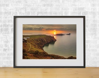 Puede incluir: Una fotografía enmarcada de una puesta de sol sobre una costa rocosa. El sol se pone detrás de una pequeña isla en la distancia, proyectando un brillo dorado sobre el agua. La costa es accidentada y verde, con un pequeño pueblo en primer plano.