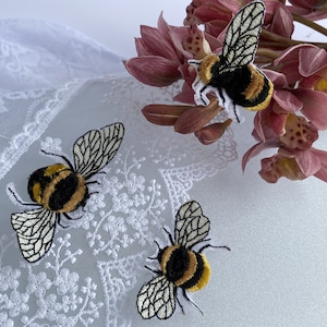 May include: Three embroidered bee patches featuring black and yellow fuzzy bodies with white wings detailed with black stitching. The patches are arranged near a piece of white lace fabric and a cluster of pink orchid flowers.