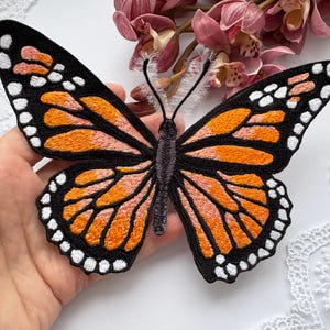 Puede incluir: Parche de mariposa bordada con detalles en naranja, negro y blanco. La mariposa tiene un patrón de alas detallado y se encuentra sobre un fondo blanco con flores rosas.