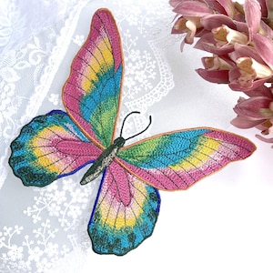 Può includere: Una toppa ricamata a forma di farfalla con vivaci sfumature di filo rosa, giallo e blu. Le ali presentano intricati motivi di cucitura con un contorno scuro e il corpo centrale è nero con due sottili antenne ricurve verso l'alto.