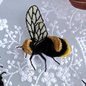 Può includere: Toppa ricamata a forma di ape nera, oro e crema. L'ape ha ali dettagliate ed è posta su uno sfondo di pizzo bianco. Questa toppa decorativa è adatta per abbigliamento o progetti artigianali.