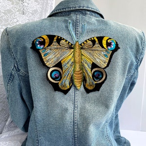Puede incluir: Una chaqueta vaquera azul con un gran parche bordado de mariposa en la espalda. La mariposa es amarilla, azul y negra con detalles intrincados.