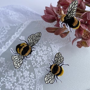 May include: Three embroidered bumblebee patches featuring black and yellow fuzzy bodies with detailed white wings with black outlines, positioned on a white lace fabric surface next to a cluster of pink orchid flowers.