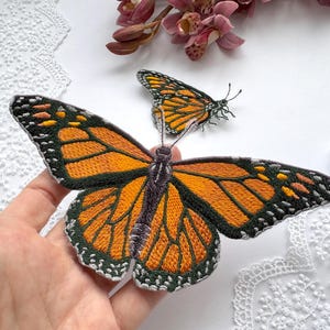 Puede incluir: Dos mariposas monarca bordadas. La mariposa más grande tiene alas naranjas y negras y un cuerpo gris. La mariposa más pequeña está en el fondo. Las mariposas están sobre una superficie blanca con un borde de encaje y flores rosas.