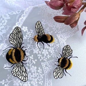 Könnte beinhalten: Drei gestickte Hummeln auf einem weißen Spitzenhintergrund. Die Bienen sind schwarz und gelb mit weißen Flügeln.