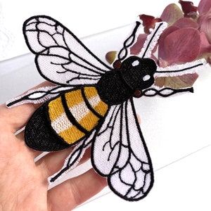 May include: An embroidered bee patch featuring a black head and thorax, a yellow and black striped abdomen, and white wings with black stitched vein details. The insect design is rendered in thread with a textured finish and defined edges.