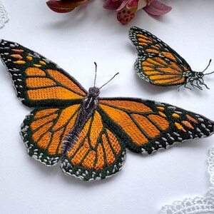 Peut inclure: Deux écussons brodés en forme de papillon monarque aux ailes orange vif avec des nervures noires, des bordures noires à points blancs et des corps gris foncé. Le plus grand papillon est à gauche, le plus petit est placé sur la droite.