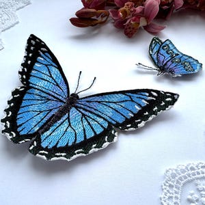 Puede incluir: Dos parches bordados de mariposas. La mariposa más grande tiene alas de color azul claro con detalles en blanco y negro. Una mariposa más pequeña está en el fondo con alas moradas y azules. Las mariposas están sobre una superficie blanca.