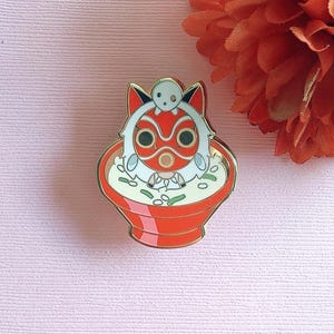 May include: A gold enamel pin depicting a red and white character design above a bowl of food. The character has a wolf-like face with a small white spirit on top. The bowl is red with white and green details.
