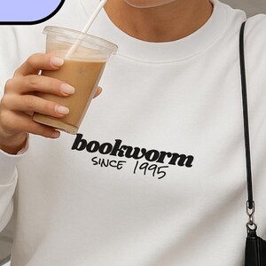 Könnte beinhalten: Weißes Sweatshirt mit dem Wort "bookworm" in Schwarz aufgedruckt, sowie "SINCE 1995". Das Sweatshirt wird mit einem weißen Rock und einer schwarzen Handtasche getragen. Das Bild zeigt auch ein Getränk in einem durchsichtigen Becher.
