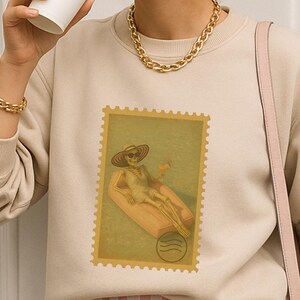 Könnte beinhalten: Beiges Sweatshirt mit einem Briefmarken-Motiv, das eine Person zeigt, die sich auf einer Schwimminsel entspannt. Die Person trägt einen Hut und eine Sonnenbrille. Das Model trägt eine Sonnenbrille, Goldschmuck und rosa-weiß gestreifte Shorts.