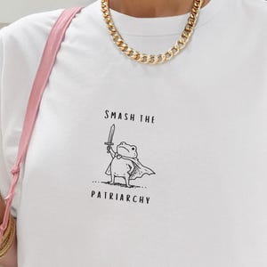 Könnte beinhalten: Weißes T-Shirt mit dem Text "SMASH THE PATRIARCHY" und einer Frosch-Illustration. Das Shirt wird mit einer rosa-weiß gestreiften Hose und Goldschmuck getragen. Eine rosa Tasche und eine Kaffeetasse sind ebenfalls zu sehen.