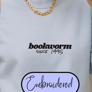 Könnte beinhalten: Hellblaues Sweatshirt mit dem Text "bookworm SINCE 1995" in Schwarz. Darunter ein hellvioletter Oval mit dem Wort "Embroidered" in Schreibschrift. Das Sweatshirt hat lange Ärmel und einen Rundhalsausschnitt.