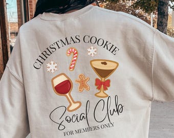 May include: A beige crewneck sweatshirt featuring a back graphic with the text "CHRISTMAS COOKIE SOCIAL CLUB FOR MEMBERS ONLY" in black, accompanied by illustrations of a wine glass, a martini glass, a candy cane, a gingerbread man, and snowflakes.
