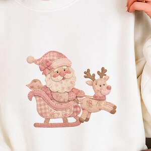 Könnte beinhalten: Cremefarbenes Sweatshirt mit einem skurrilen Weihnachtsmann- und Rentier-Design. Der Weihnachtsmann ist in einem rosa karierten Outfit dargestellt und sitzt in einem Schlitten, der von einem Rentier gezogen wird. Das Design ist in weichen, gedämpften Farben gehalten.