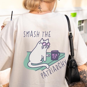 May include: Cream-colored t-shirt with the text "SMASH THE PATRIARCHY" and a graphic of a white cat wearing glasses, sitting next to a mug. The cat is on a green surface. The shirt is a casual, oversized fit.