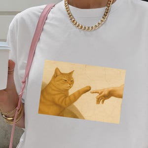 Könnte beinhalten: Ein weißes Kurzarm-T-Shirt mit einem rechteckigen Grafikdruck, der eine orangefarbene getigerte Katze zeigt, die ihre Pfote nach einem menschlichen Finger ausstreckt, im Stil der Erschaffung Adams auf beigem Hintergrund.