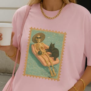 Op de afbeelding: Lichtroze T-shirt met een postzegel-achtig ontwerp van een skelet en een zwarte kat op een zwemband. Het model draagt een roze en wit gestreepte short en houdt een roze tas en een witte koffiebeker vast.