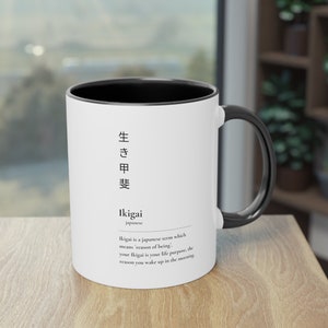 Könnte beinhalten: Eine weiße Keramiktasse mit schwarzem Rand und Henkel. Die Tasse zeigt das japanische Wort "生き甲斐" (Ikigai) und eine Definition des Wortes auf Englisch. Die Definition lautet: "Ikigai ist ein japanischer Begriff, der 'Grund des Seins' bedeutet. Dein Ikigai ist dein Lebenszweck, der Grund, warum du morgens aufwachst."