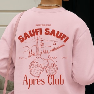 Op de afbeelding: Een lichtroze sweatshirt met een rood grafisch ontwerp op de achterkant. Het ontwerp bevat de woorden "SAUFI SAUFI" boven een berglandschap met skiliften en de woorden "Apres Club" eronder. Het sweatshirt heeft ook de tekst "Sincerely Yours Presents" en "EST. 2025".