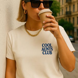 Könnte beinhalten: Ein cremefarbenes Kurzarm-T-Shirt mit dem Text COOL AUNTS CLUB, der in fetten blauen Buchstaben auf der Brust gedruckt ist. Das Shirt hat einen klassischen Rundhalsausschnitt und einen geraden Saum für einen schlichten Look.
