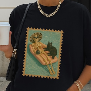 Op de afbeelding: Zwart T-shirt met een vintage postzegelontwerp. De postzegel toont een skelet op een zwemband met een zwarte kat. Het skelet draagt een zonnebril en een strohoed. Het shirt heeft een relaxte, oversized pasvorm.