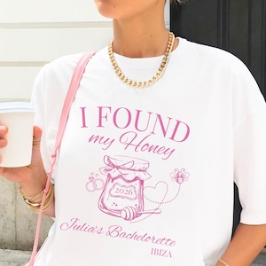 Könnte beinhalten: Ein weißes T-Shirt mit einer rosa Grafik eines Honigglases und dem Text I FOUND my Honey, 2026, Julia's Bachelorette und IBIZA. Das Design enthält einen Honiglöffel, eine kleine Biene und zwei ineinander verschlungene Ringe in Rosa.