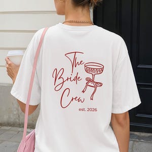 May include: A white t-shirt with the text "The Bride Crew" in a burgundy script font, along with a matching graphic of a cocktail glass and the year "est. 2026". The shirt is part of a bridal party theme.