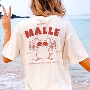 Può includere: Una t-shirt color bianco sporco con una grafica rossa sul retro. Il design include il testo MALLE, EST. 2026 e EIGENTLICH SIND WIR DOCH ZUM SAUFEN DA. Al centro è raffigurato un topo dei cartoni animati con occhiali da sole e due boccali.