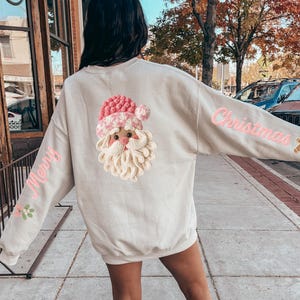 Może przedstawiać: Beżowa bluza z nadrukiem Świętego Mikołaja na plecach. Wzór Świętego Mikołaja ma różowo-białą czapkę, białą brodę i różowy nos. Na rękawach różowe napisy "Merry" i "Christmas".