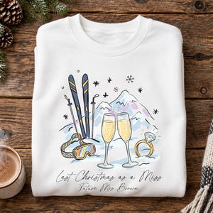 Könnte beinhalten: Ein weißes Sweatshirt mit Rundhalsausschnitt und einer grafischen Illustration von Skiern, Skibrille, zwei Champagnergläsern und einem Diamantring vor einer Bergkulisse. Der Text lautet, Last Christmas as a Miss, Future Mrs. Brown.