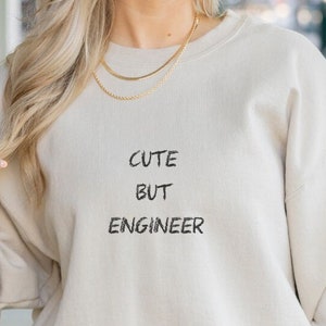 Könnte beinhalten: Ein beiger Pullover mit dem Aufdruck "Cute But Engineer" auf der Vorderseite.