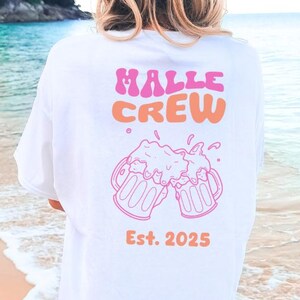 May include: White t-shirt with the text "HALLE CREW" in pink and orange, along with a graphic of two beer mugs clinking. Below the graphic is "Est. 2025" in orange. The shirt is worn on a beach.