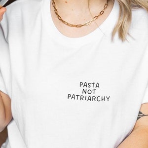 May include: A white short-sleeved t-shirt featuring the black text PASTA NOT PATRIARCHY printed on the upper left chest area. The garment has a round neckline and rolled sleeves, constructed from a plain white fabric with a simple design.