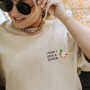 May include: A beige short-sleeved t-shirt featuring a small graphic of a mallard duck on the left chest area with the text "I DON'T GIVE A QUACK." printed in black capital letters next to the duck illustration.